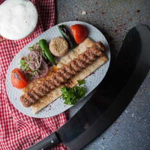 Top view adana kebab with knife and fried vegetables and chopped onion and ayran in white plate on stone background