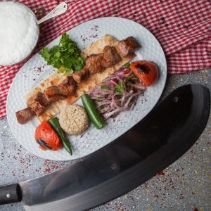 Top view shish kebab with fried vegetables and chopped onion and ayran and knife in white plate on stone background